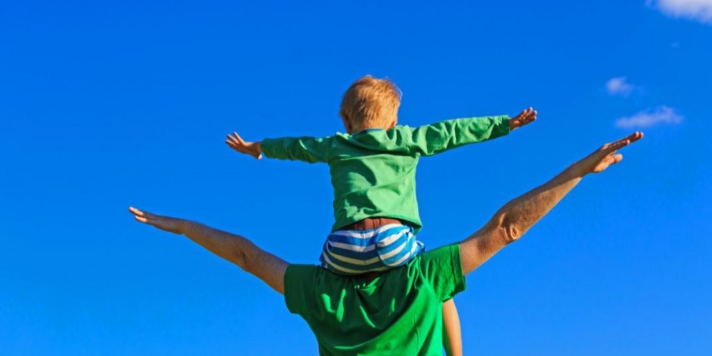 Zoontje zit op de schouders van zijn papa. Beide hebben zijdelings gestrekte armen.
